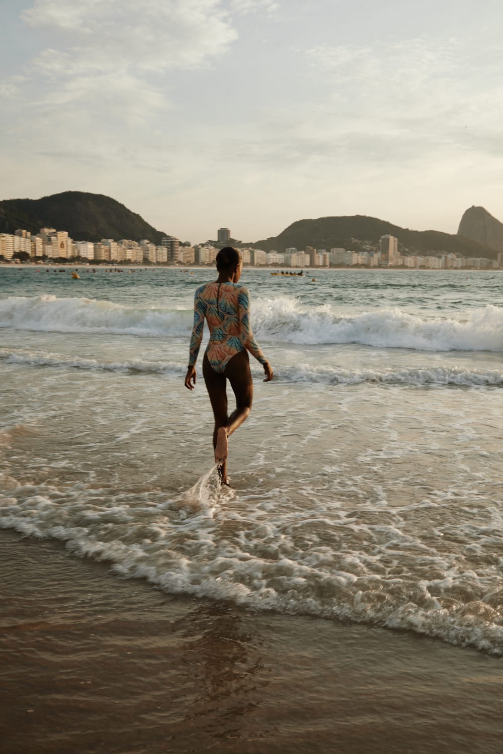 Modelo caminhando na praia de Copacabana com maiô de estampa exclusiva da Ph Praia, Pão de Açúcar ao fundo